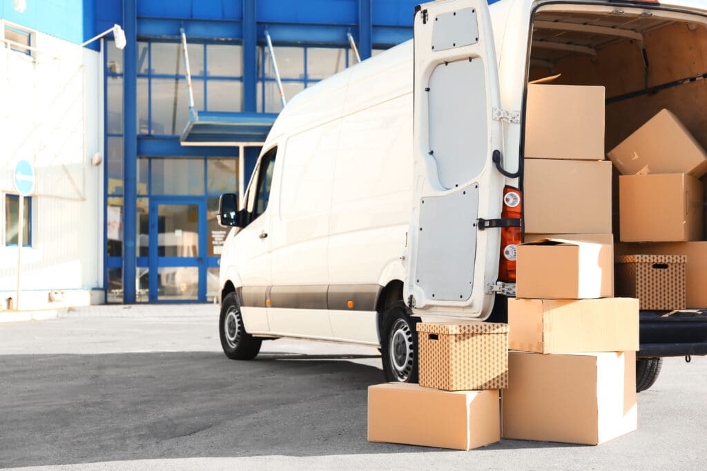 A white delivery van, marked for Same Day Courier service, is parked outside a building with its back doors open and several cardboard boxes stacked on the ground and inside the van, ready for urgent delivery or loading.