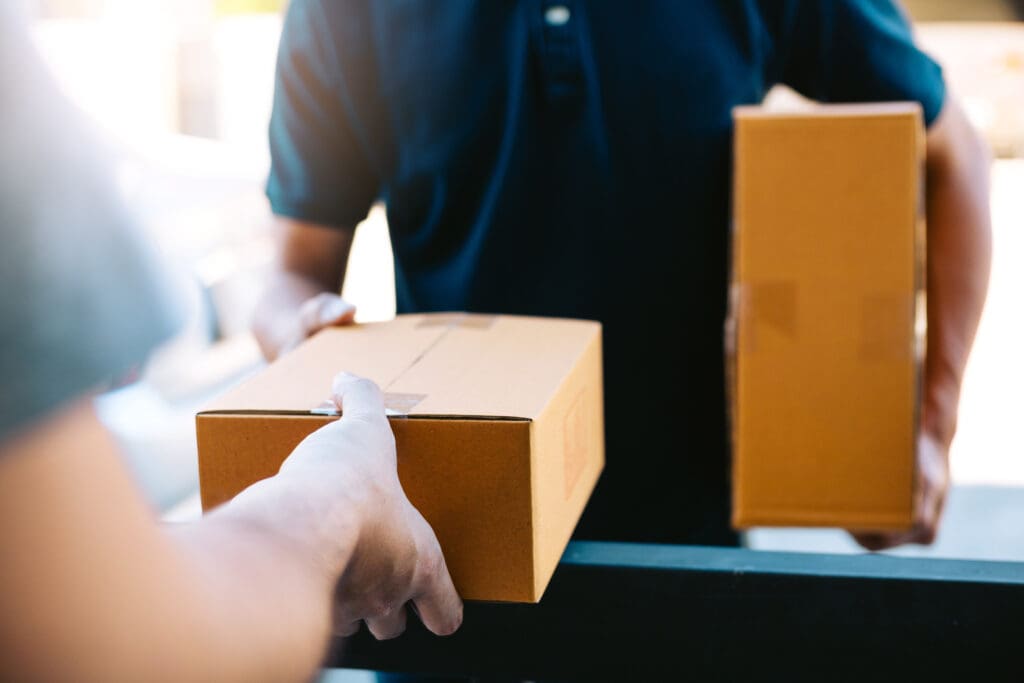 A person hands a brown cardboard box to another individual while holding a second box, suggesting an urgent delivery or package exchange by a same day courier. Both people's faces are out of view.