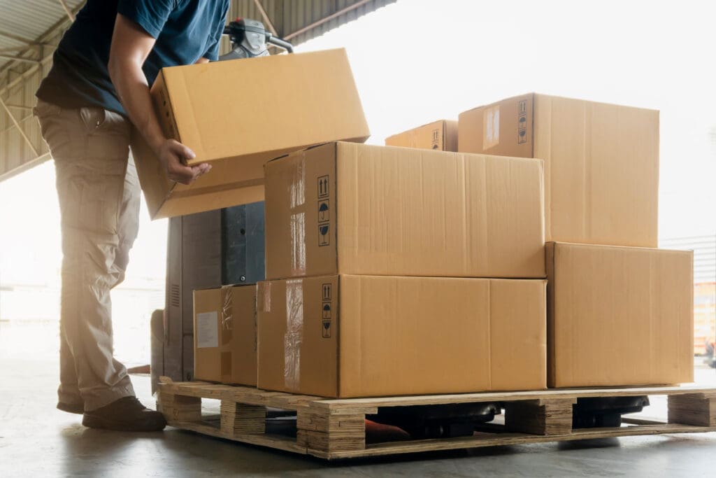 A person wearing beige pants and a navy shirt is loading or unloading cardboard boxes onto a wooden pallet inside a warehouse, ensuring efficient logistics for urgent delivery services.