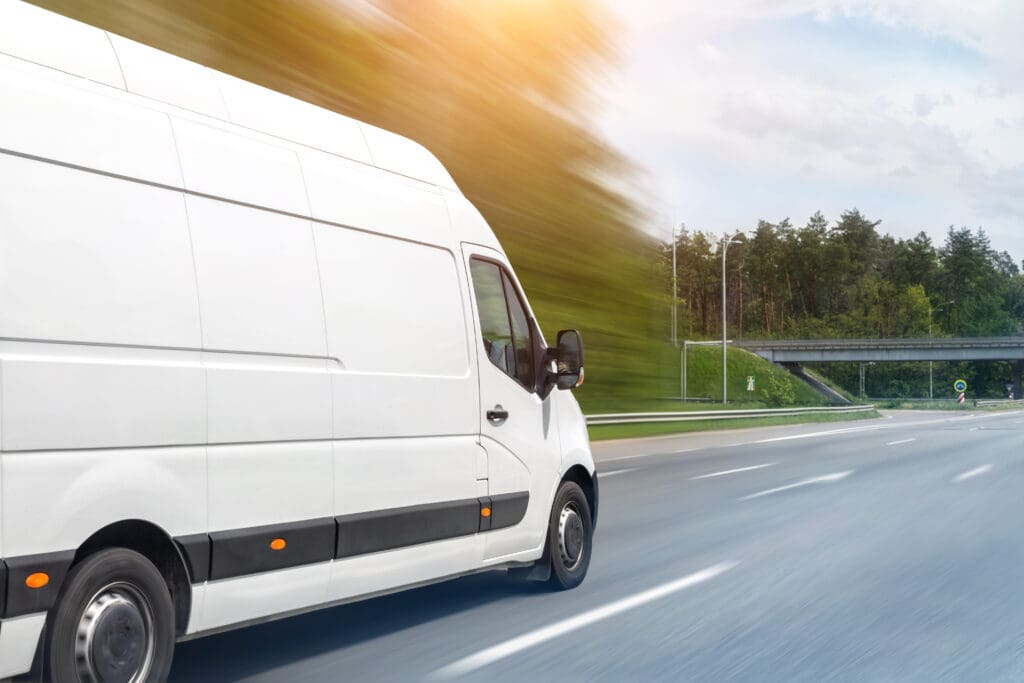 A white Same Day Courier van drives quickly down a highway, with blurred trees and sunlight in the background, suggesting urgent delivery and speed on a clear day.