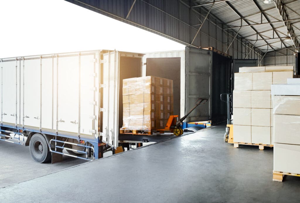 A warehouse loading dock with trucks being loaded for urgent delivery, using a pallet jack to move pallets of cardboard boxes. Stacks of boxes are ready for shipment in a well-lit industrial logistics setting.