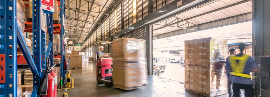 Busy warehouse with high shelves, stacks of cardboard boxes on pallets, a worker in a safety vest, and a forklift moving goods for urgent delivery near an open loading dock in daylight.